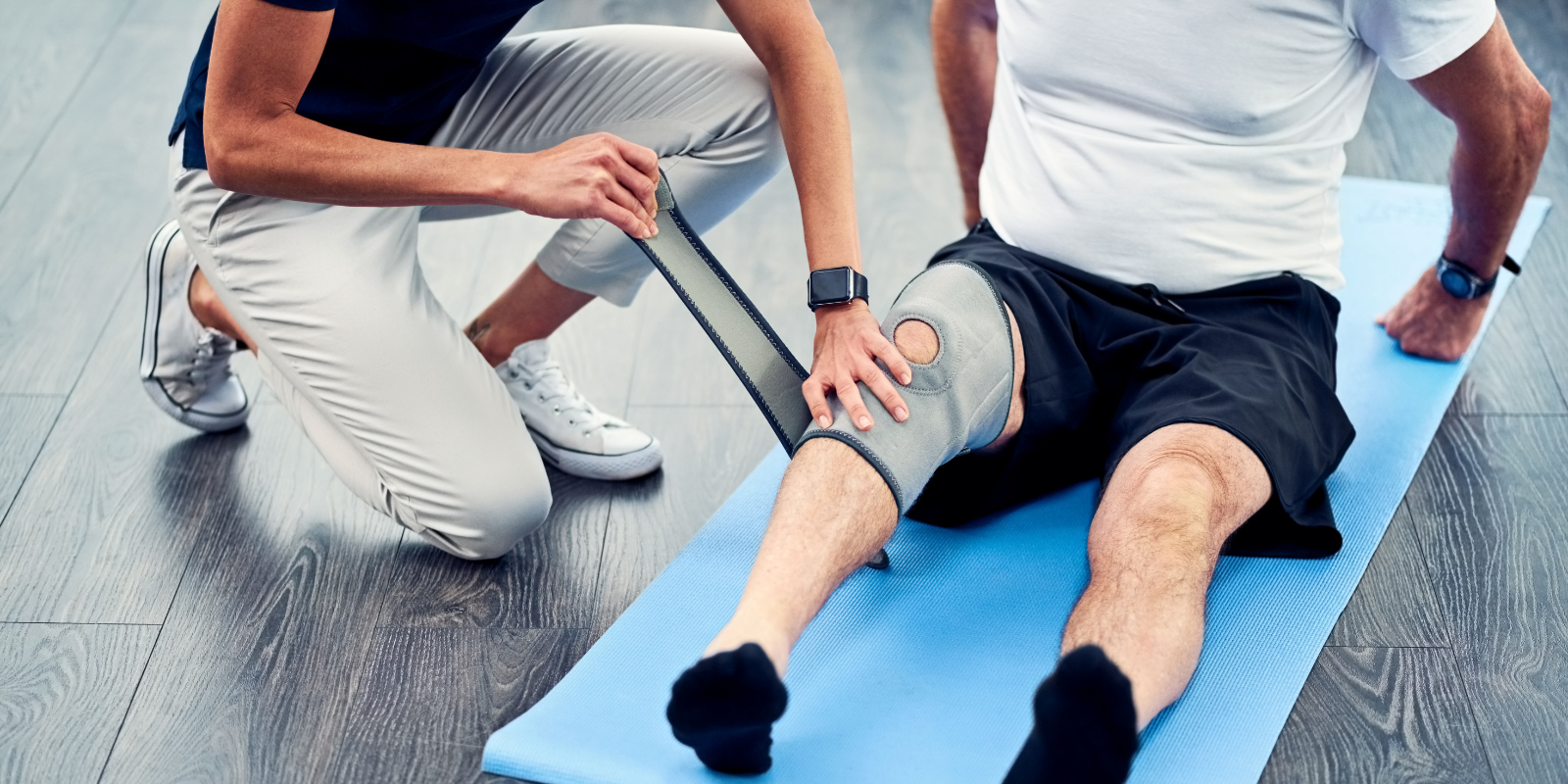 Woman putting knee brace on man on yoga mat during rehabilitation